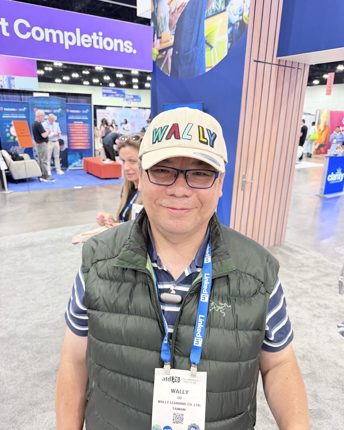Smiling attendee on a trade show floor wearing a cream cap personalized with his name in multicolor lettering